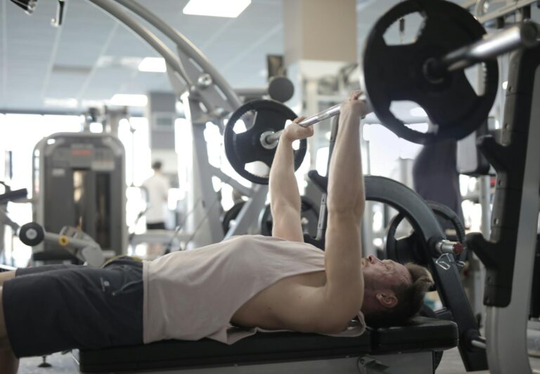 Side view of strong young sportsman in casual sportswear pressing heavy barbell on bench while intense training in modern fitness center