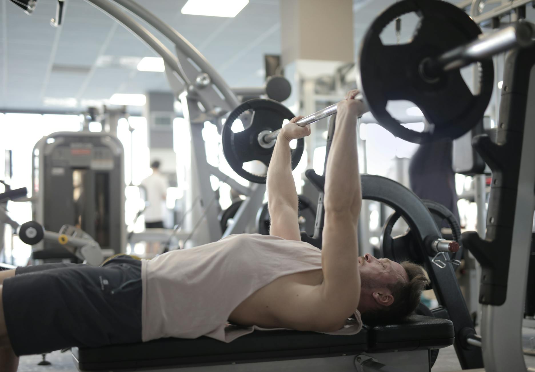 Side view of strong young sportsman in casual sportswear pressing heavy barbell on bench while intense training in modern fitness center