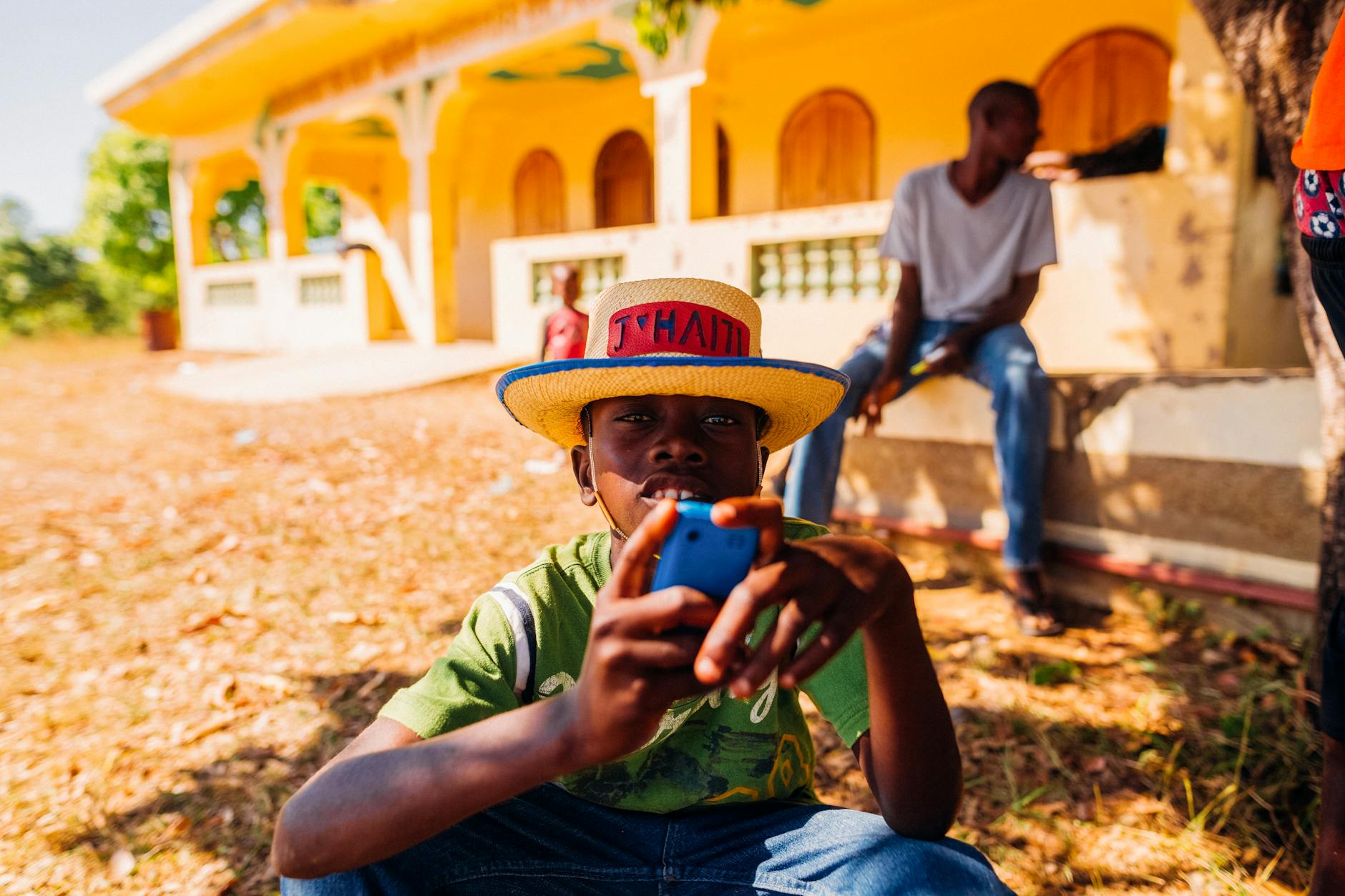 Concentrated African boy in casual clothes and straw hat using mobile phone while resting on yard near house in village on sunny day