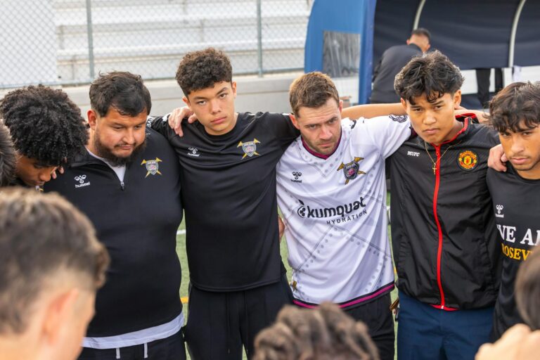 A soccer team group huddle shows unity and teamwork on an outdoor field.