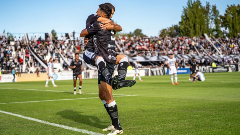 Two soccer players joyfully celebrate a goal during a match on a sunny day.
