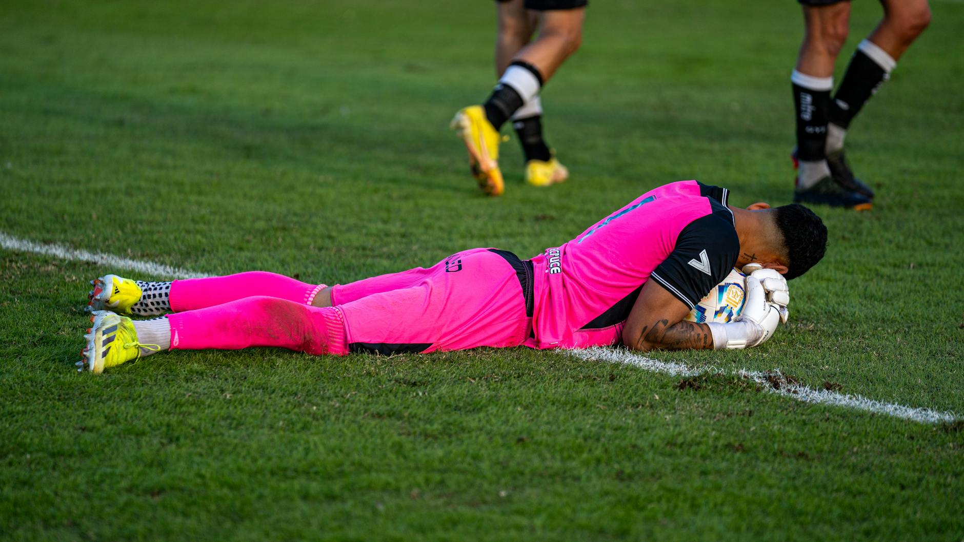 Soccer goalie in bright pink kit lying on grass field during a match.