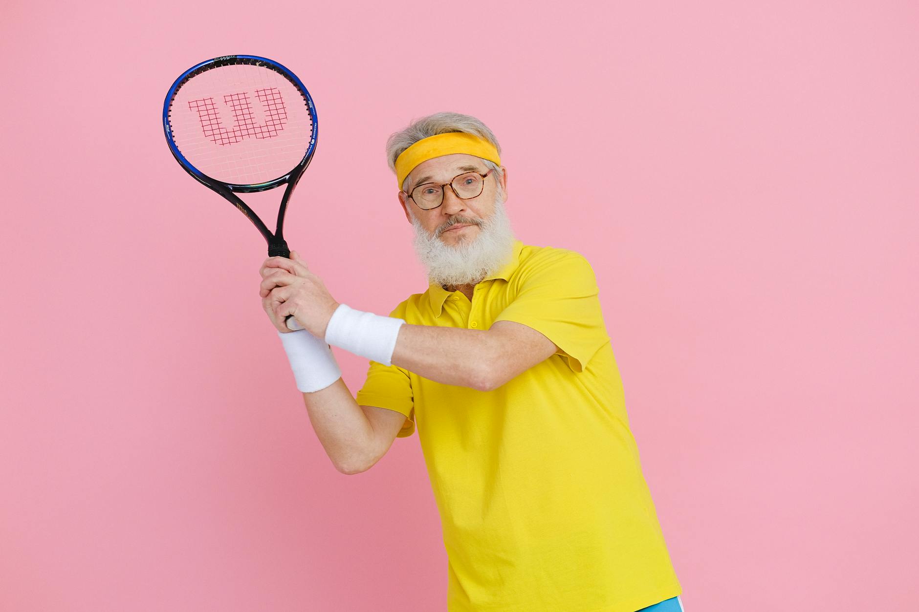 Elderly man in yellow sportswear holding a tennis racket against a vibrant pink background.