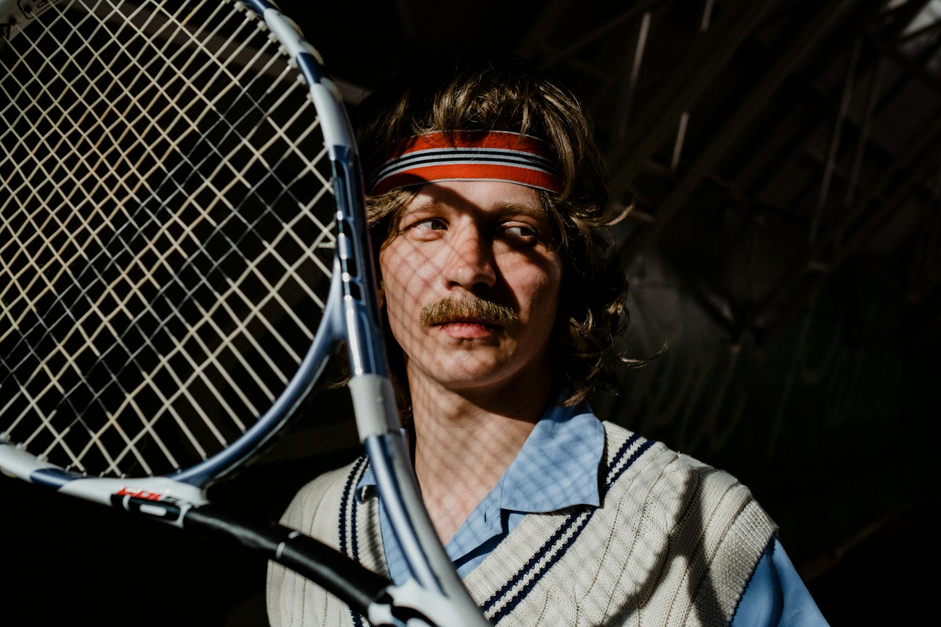 Vintage-style portrait of a man with a mustache holding a tennis racket indoors.