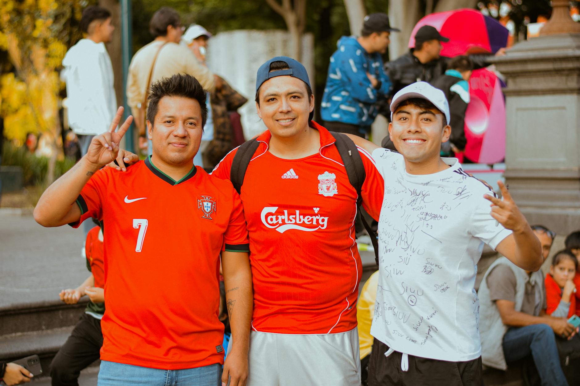 Three young men smiling and posing in sports jerseys outdoors, showing peace signs.
