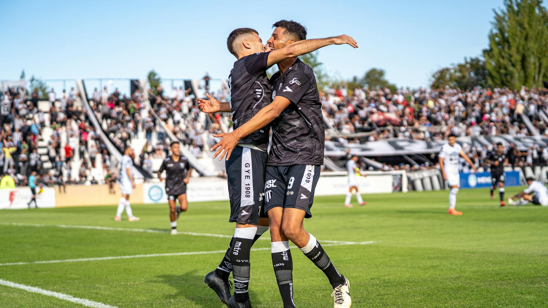 Two soccer players in black jerseys celebrate a goal on a sunny day at a packed stadium.