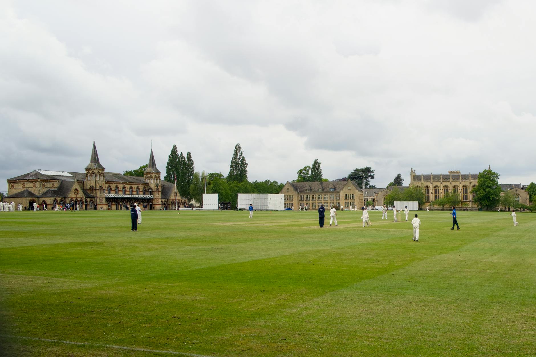 Outdoor cricket match on a lush green field with a historic building backdrop, overcast sky, many players visible.