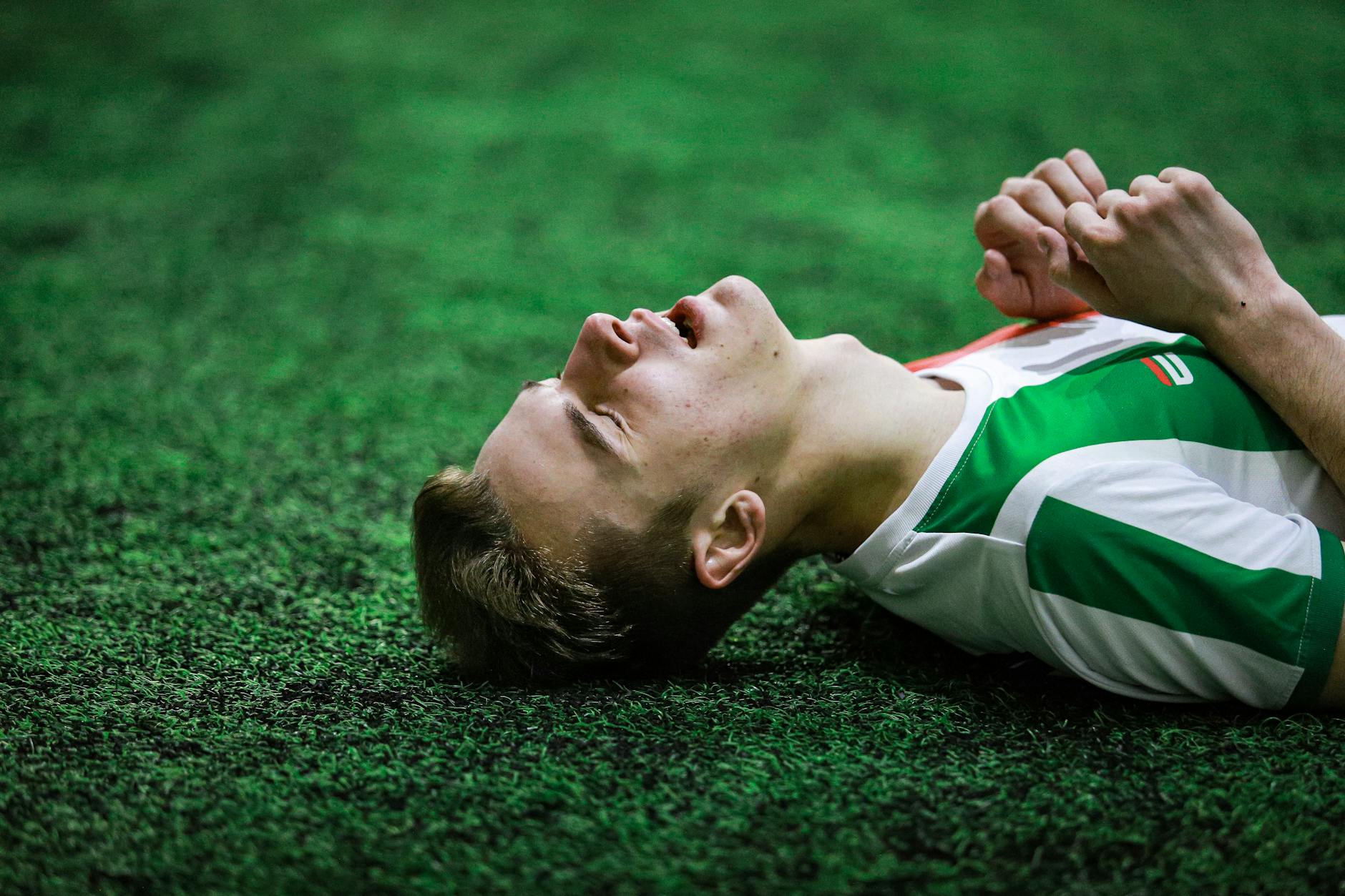 Young male soccer player in green jersey lying on grass, eyes closed, exhausted.