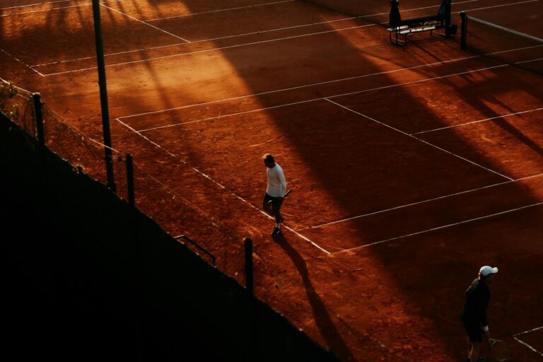 Dynamic aerial shot of a tennis match on a clay court during sunset, capturing shadows and movement.