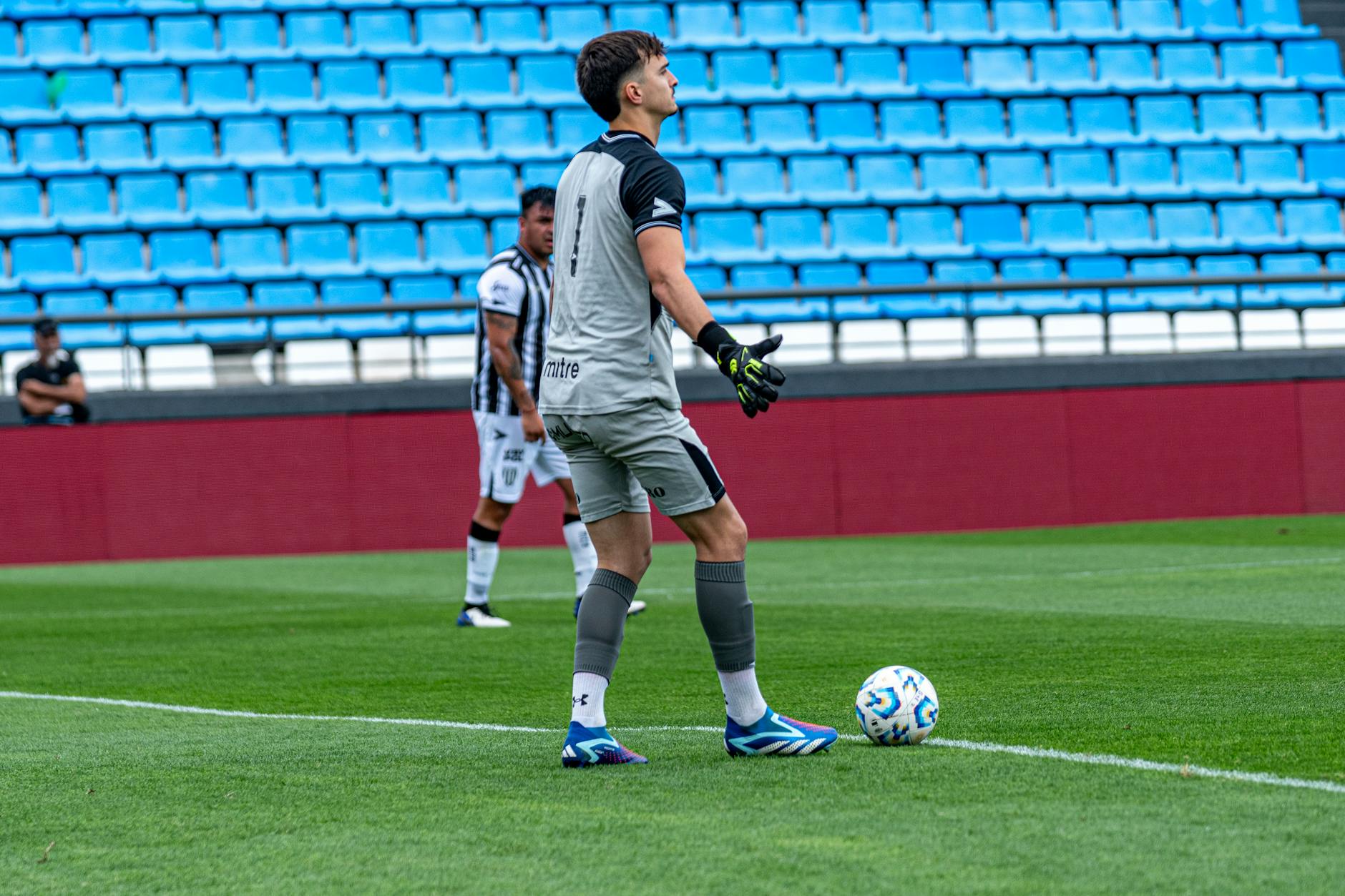 Goalkeeper holding a soccer ball during a match on a bright green field with empty blue seats in the background.