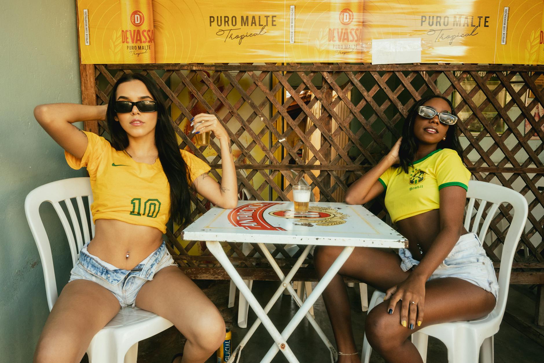 Two fashionable women in Brazil jerseys relax at an outdoor cafe table, showcasing modern summer style.