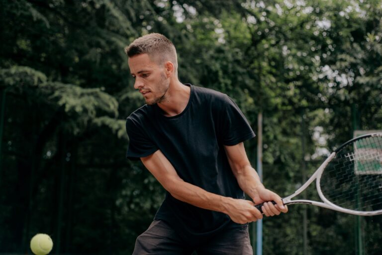 Caucasian man in black shirt playing tennis outdoors, preparing to hit the ball with determined focus.