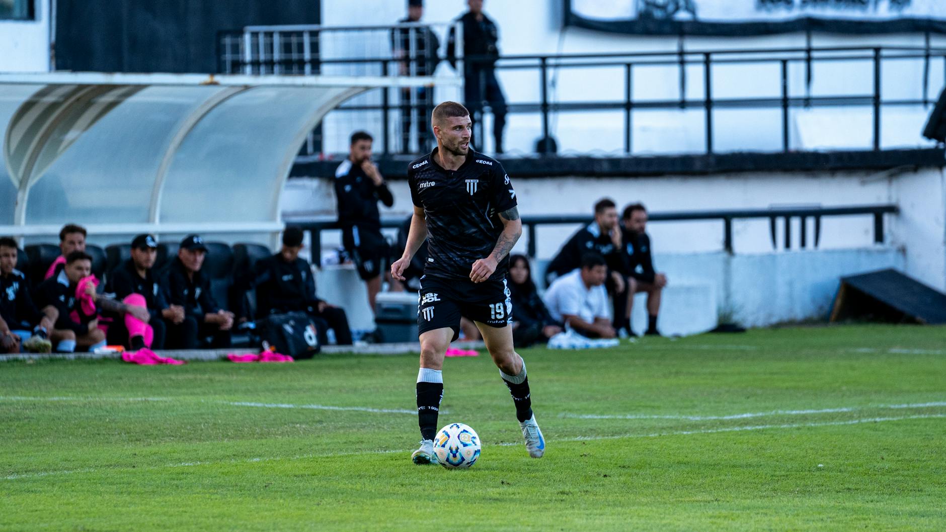 Soccer player dribbling during a match with team watching from the sidelines.