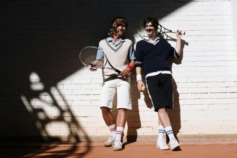 Two friends wearing vintage tennis attire smile playfully on a sunlit court.