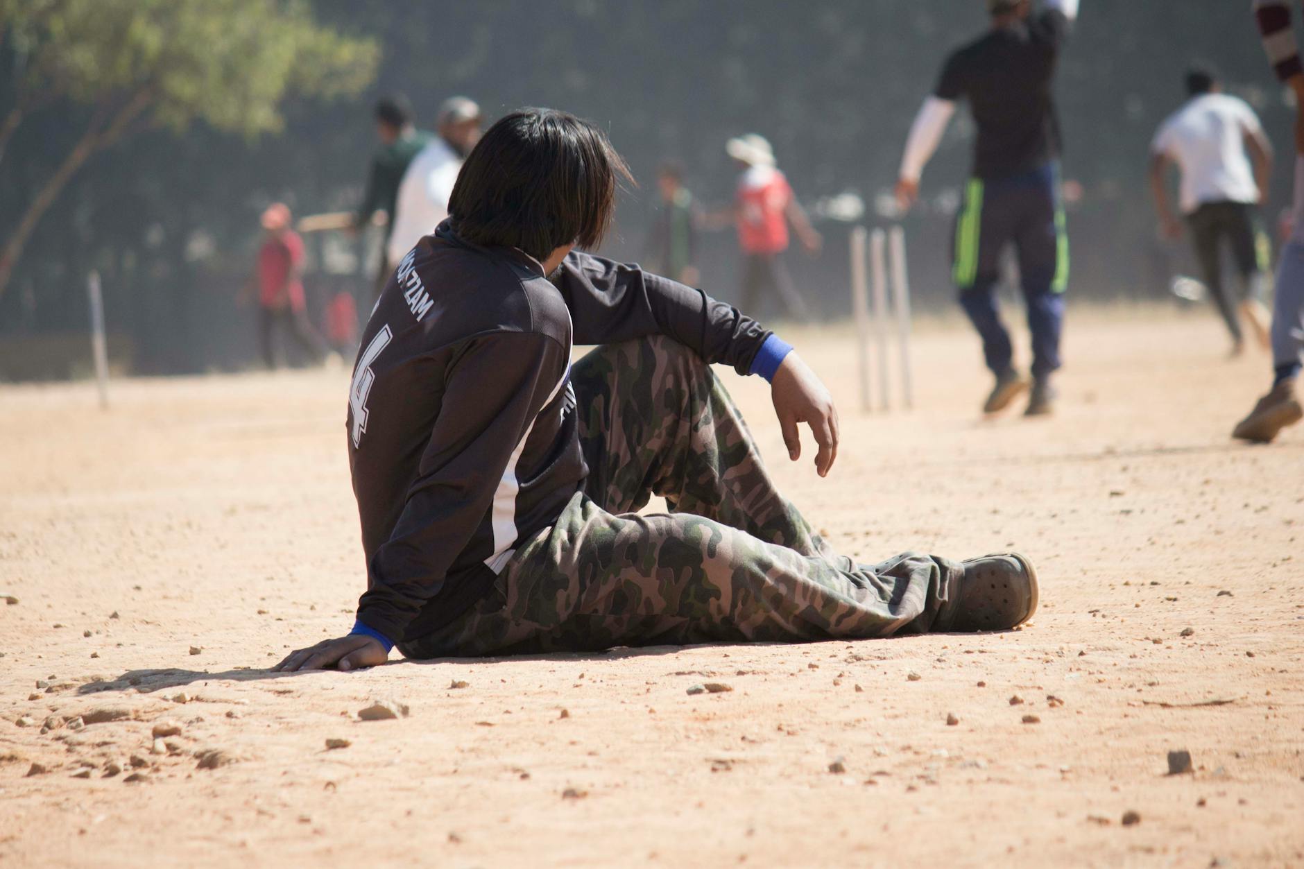 A young man sits on a dusty cricket field in the sun, wearing a jersey and camouflage pants.