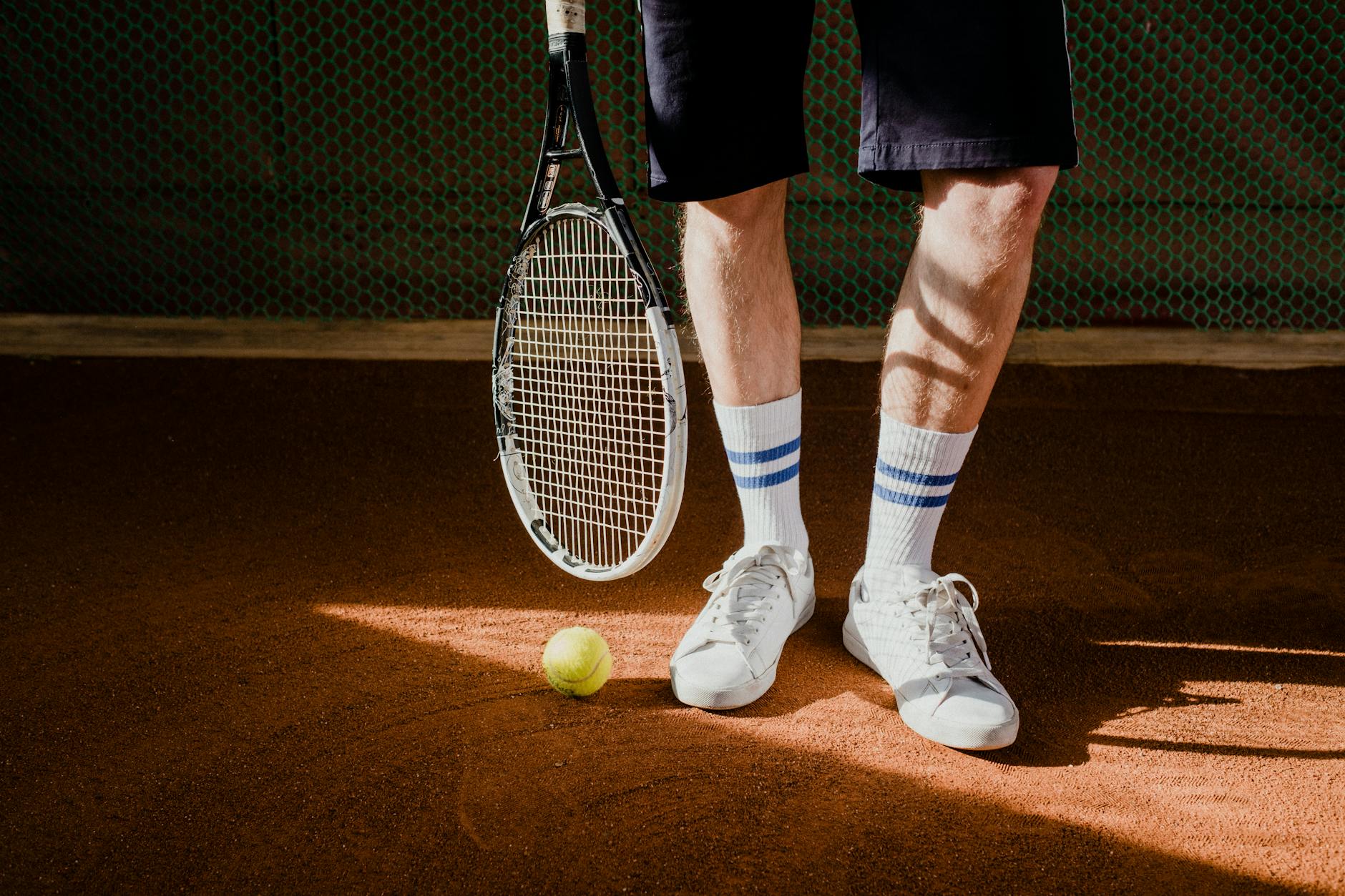 Close-up of a tennis player with racket and ball on a clay court, wearing white sneakers and socks.