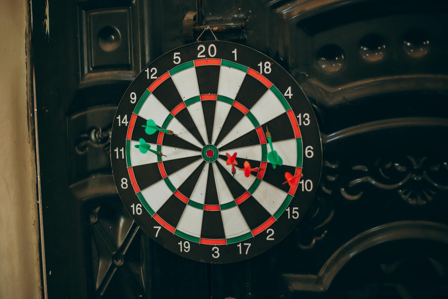 Close-up of a dartboard with red and green darts hitting various spots, indoors setting.