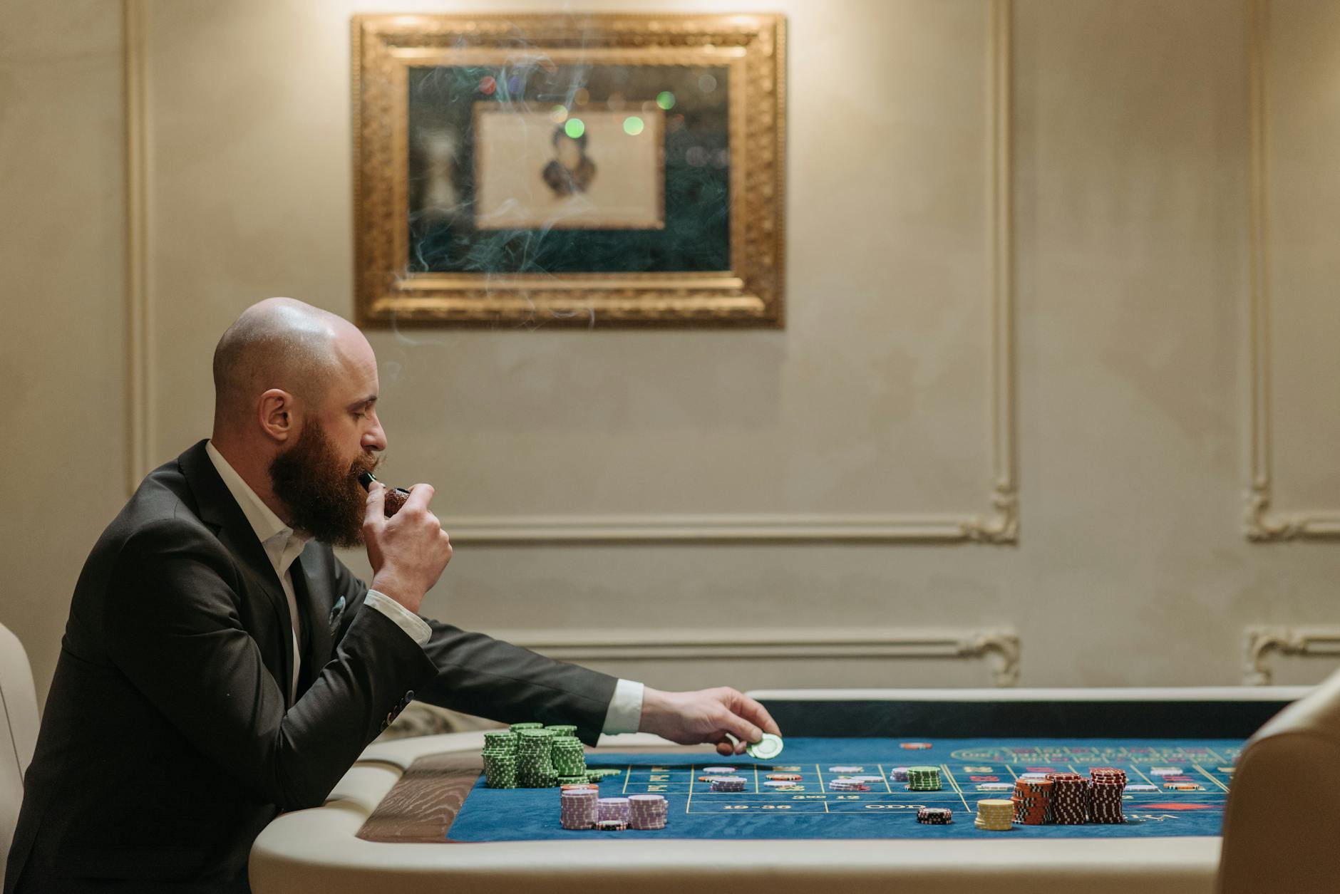 A man in a suit smokes a pipe while playing with poker chips at a casino table indoors.