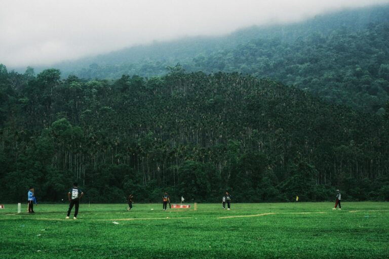 Cricketers playing in a lush green field surrounded by a forest in Sylhet, Bangladesh.