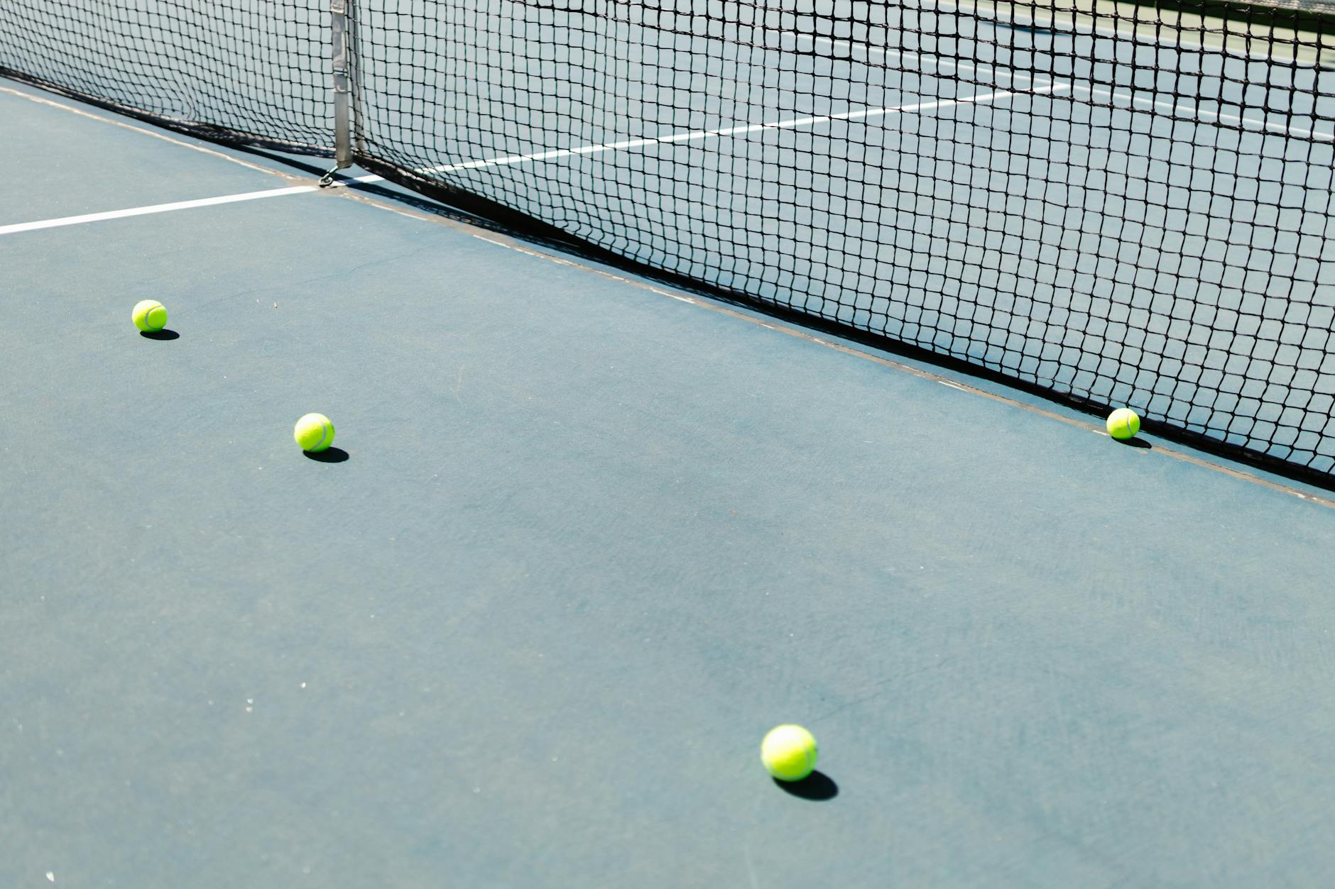 A tennis court featuring a net and several tennis balls lying on the ground, well-lit by sunlight.