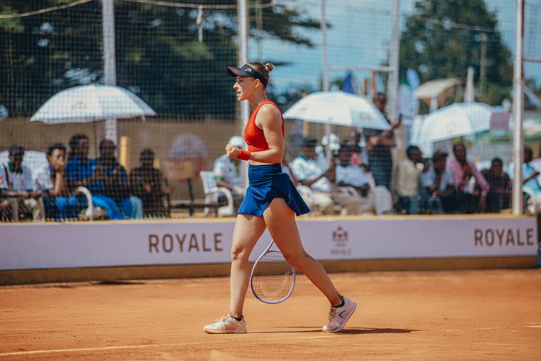 A triumphant female tennis player in action on a clay court with spectators in the background.