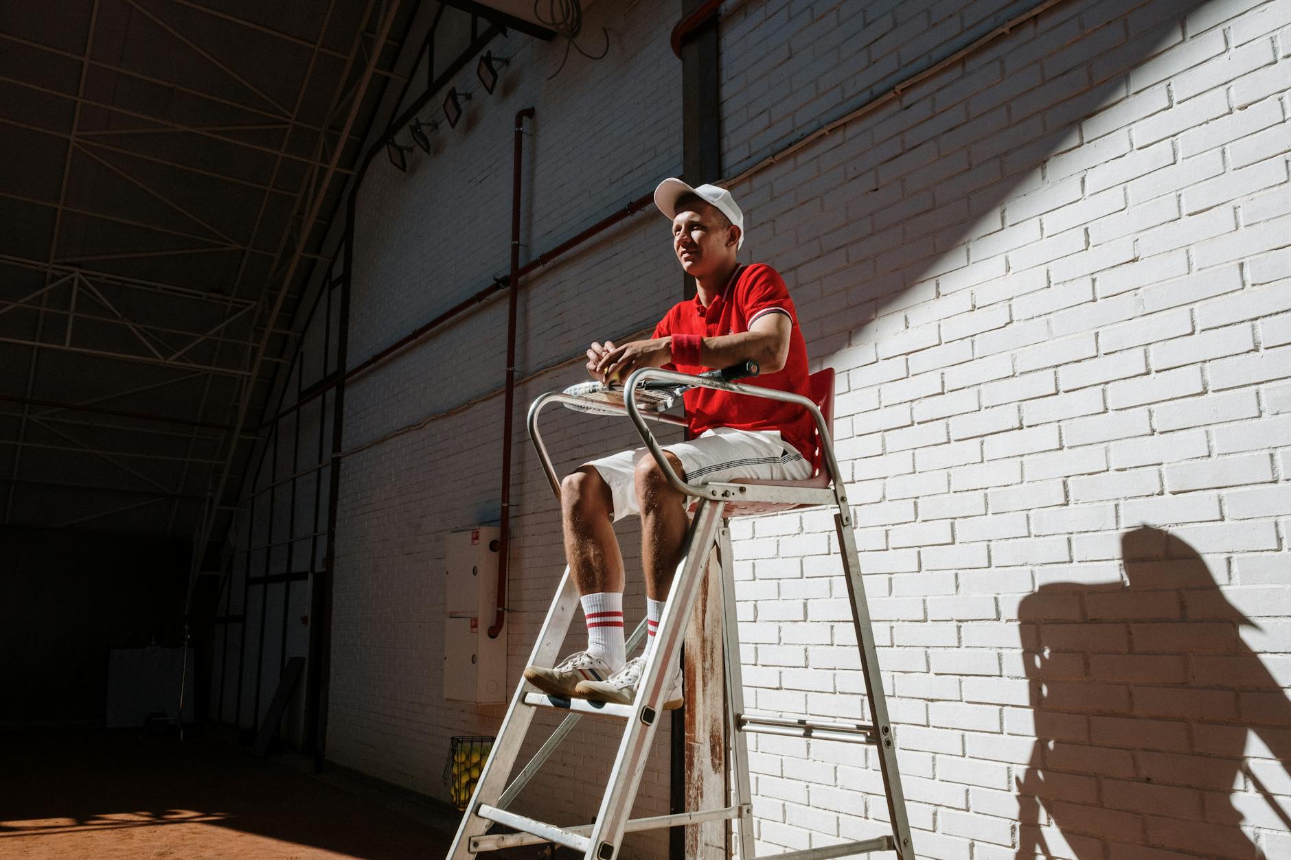 Tennis umpire sitting on a high chair against a white brick wall, creating dynamic shadows indoors.