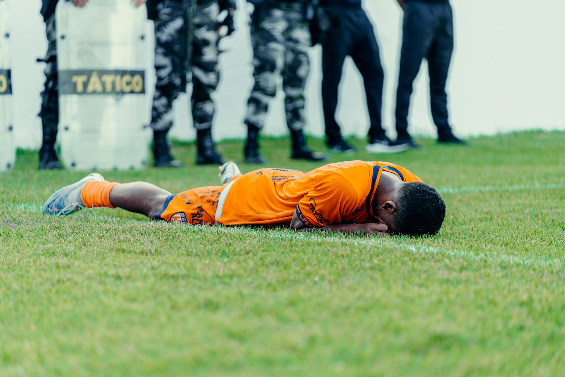 Injured football player in orange jersey lying on field with police presence.
