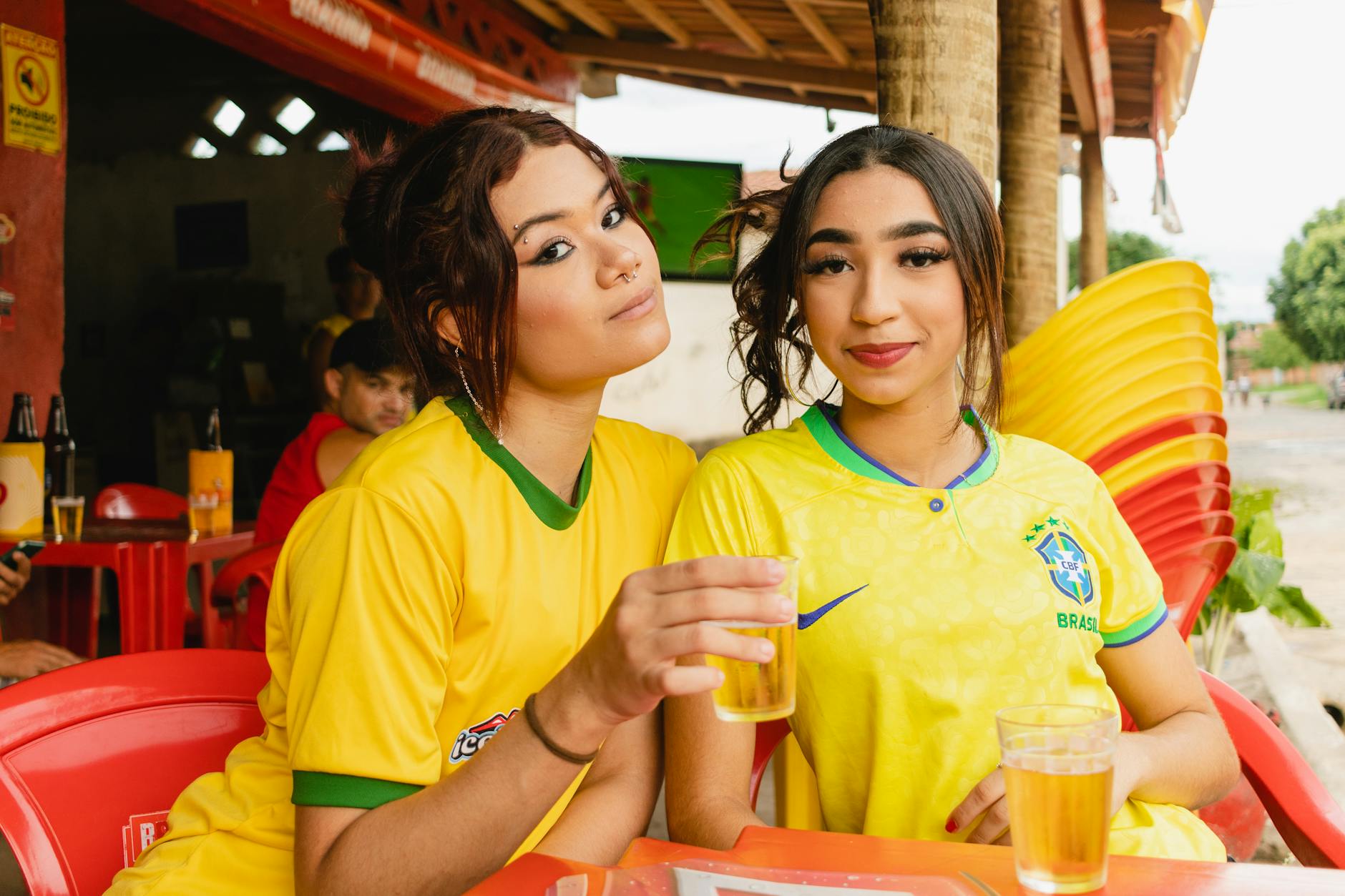 Two women in Brazil jerseys enjoying drinks at an outdoor venue.