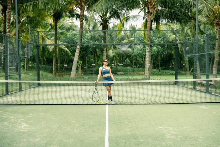 A woman poses with a tennis racket on a palm tree-lined tennis court outdoors.