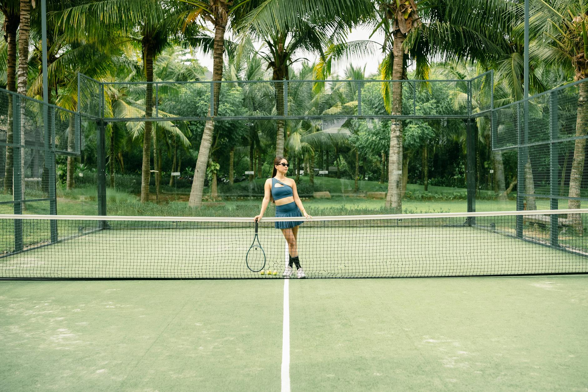 A woman poses with a tennis racket on a palm tree-lined tennis court outdoors.
