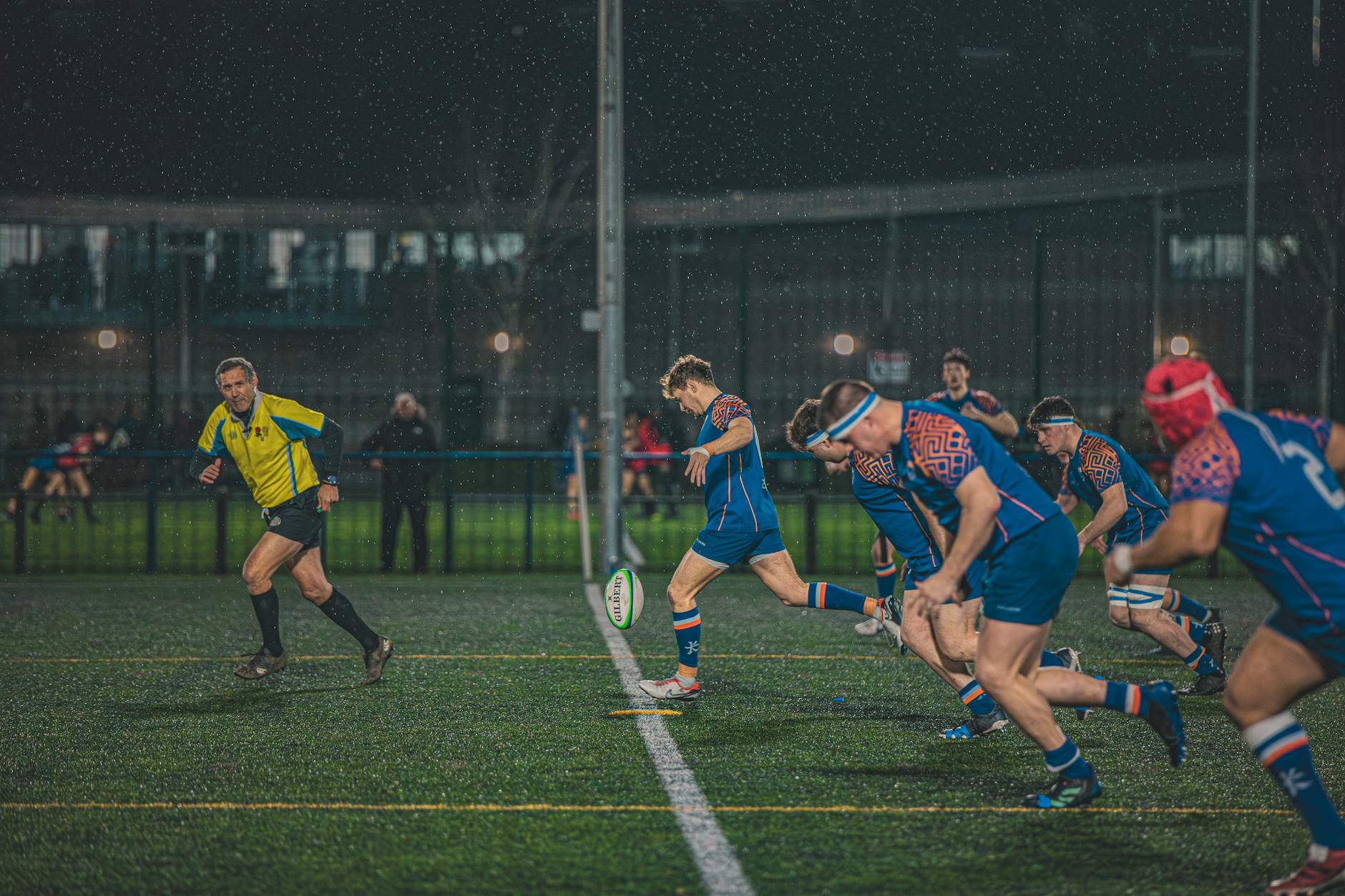 Intense night-time rugby match in England, capturing action and teamwork on a rainy field.