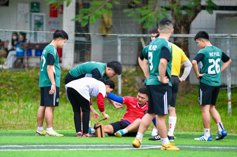 Teammates help an injured player during a soccer match on an outdoor field in Hà Nội, Việt Nam.