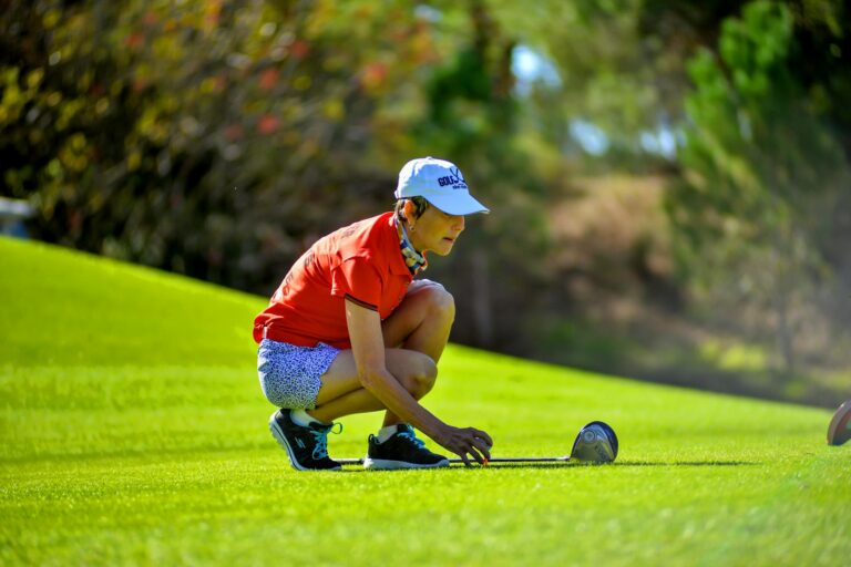 Female golfer in red prepares to putt on a lush green course under clear skies.