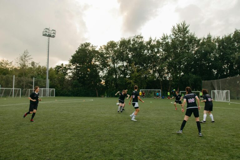 Women having an outdoor soccer game on a grassy field surrounded by trees.