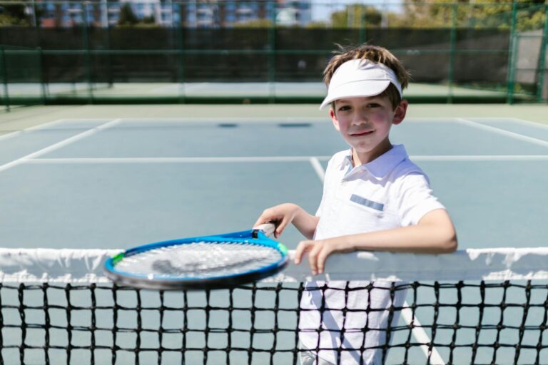 A smiling young boy in sportswear holding a tennis racket on a sunny outdoor court.