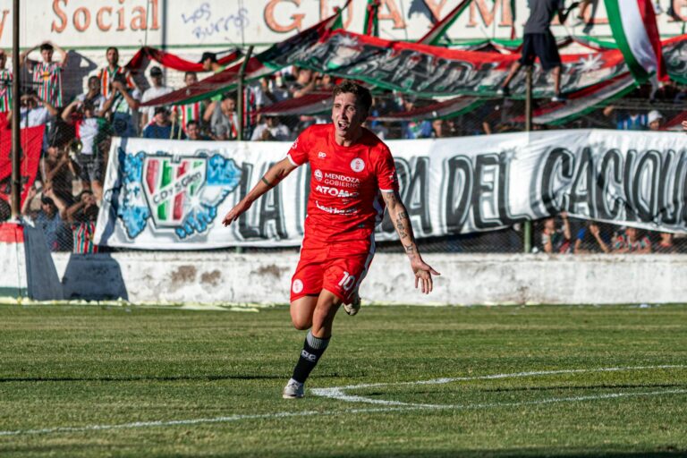 Soccer player in red uniform celebrating a goal on the field with cheering fans in the background.