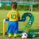 A young child wearing a Neymar Jr. jersey stands by a mini soccer goal on a grassy field.
