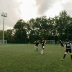 Women having an outdoor soccer game on a grassy field surrounded by trees.