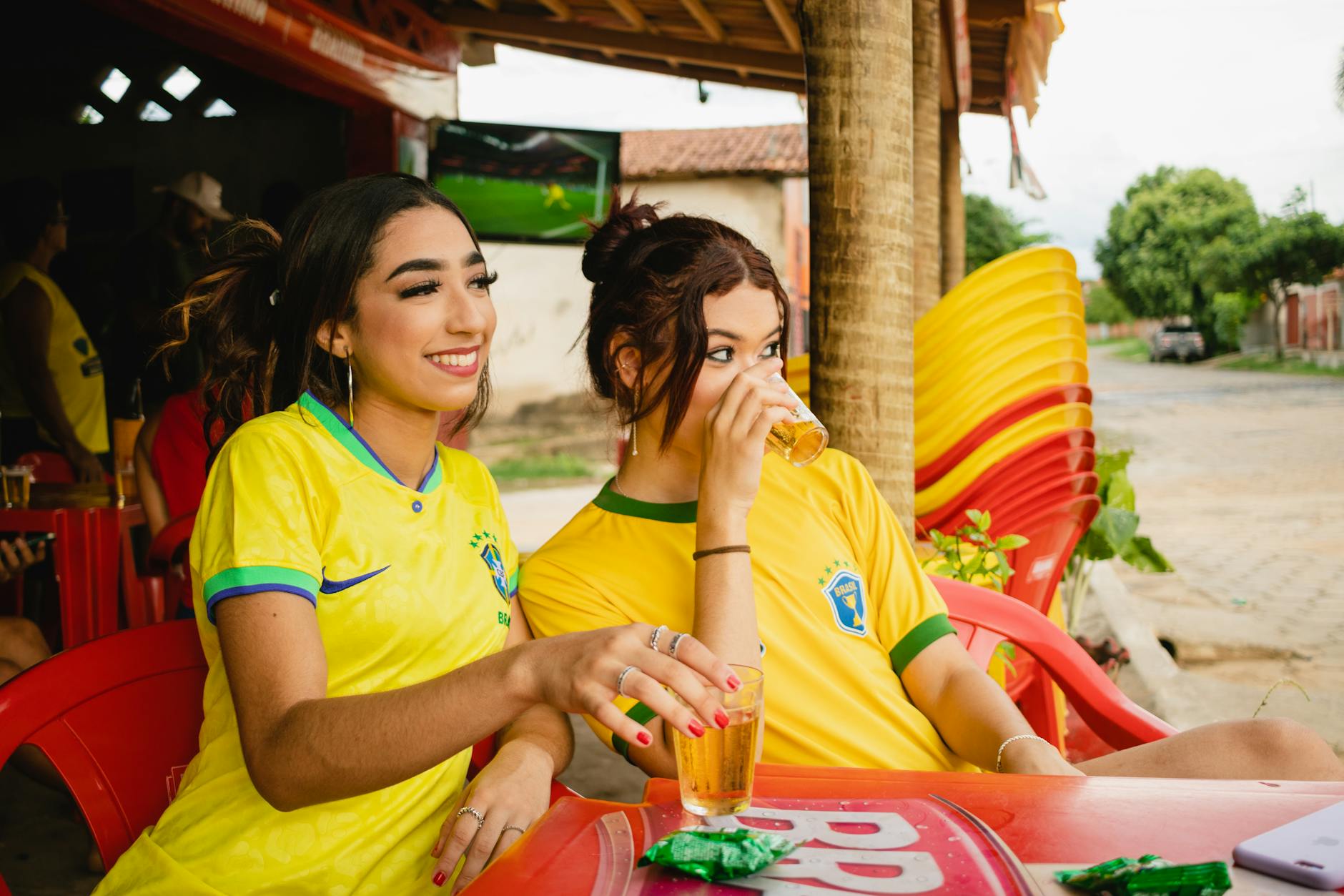 Two young women in Brazil jerseys enjoying drinks at an outdoor café, capturing summer leisure.