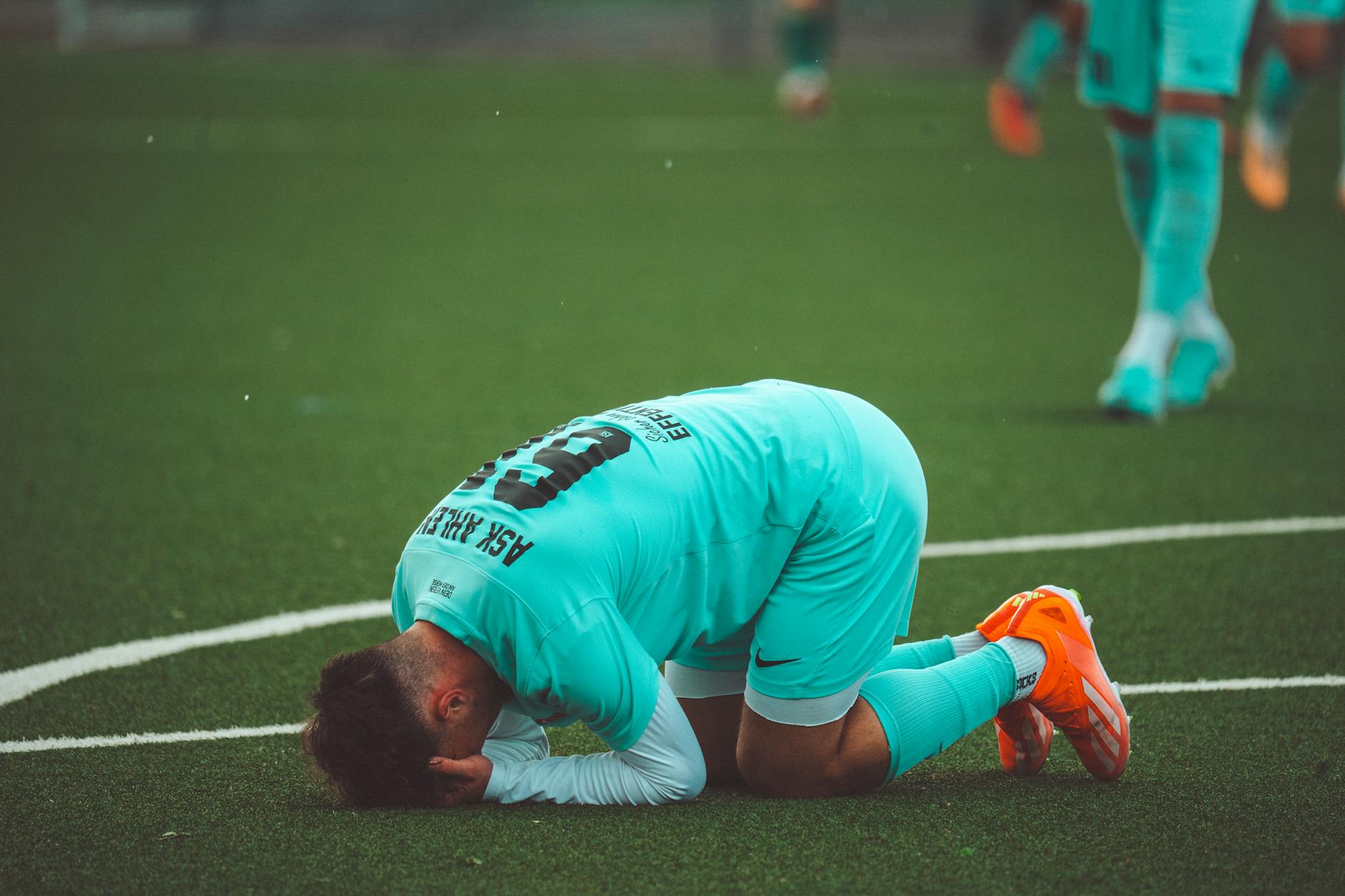 A soccer player kneels in despair on a green artificial turf sports field.