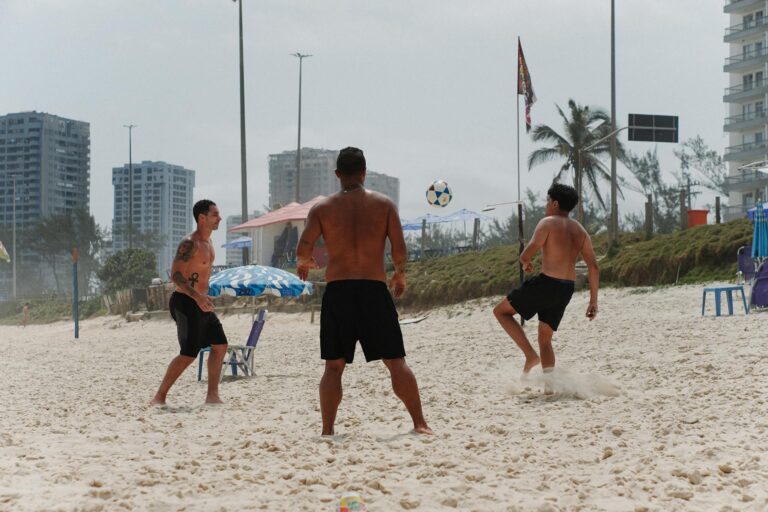 Three men playing soccer on a sandy beach with city skyline and palm trees.
