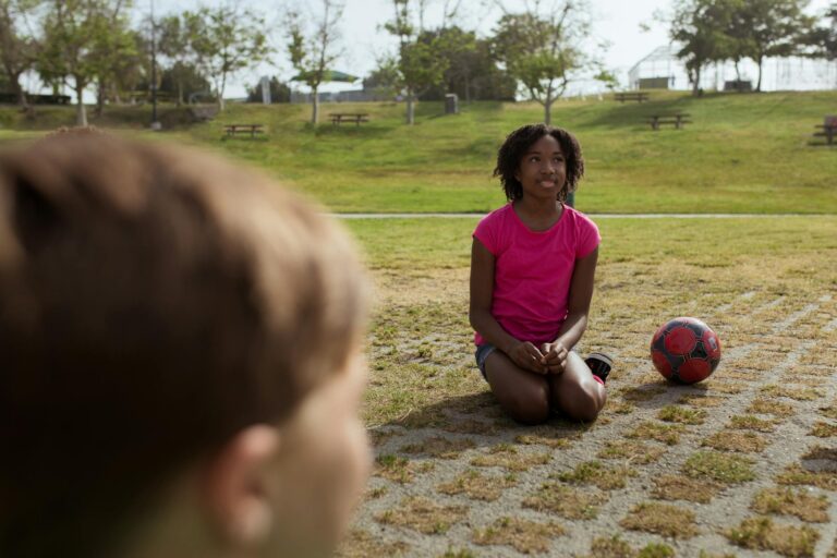 A girl sitting on grass beside a soccer ball in a sunny park, enjoying a playful day.