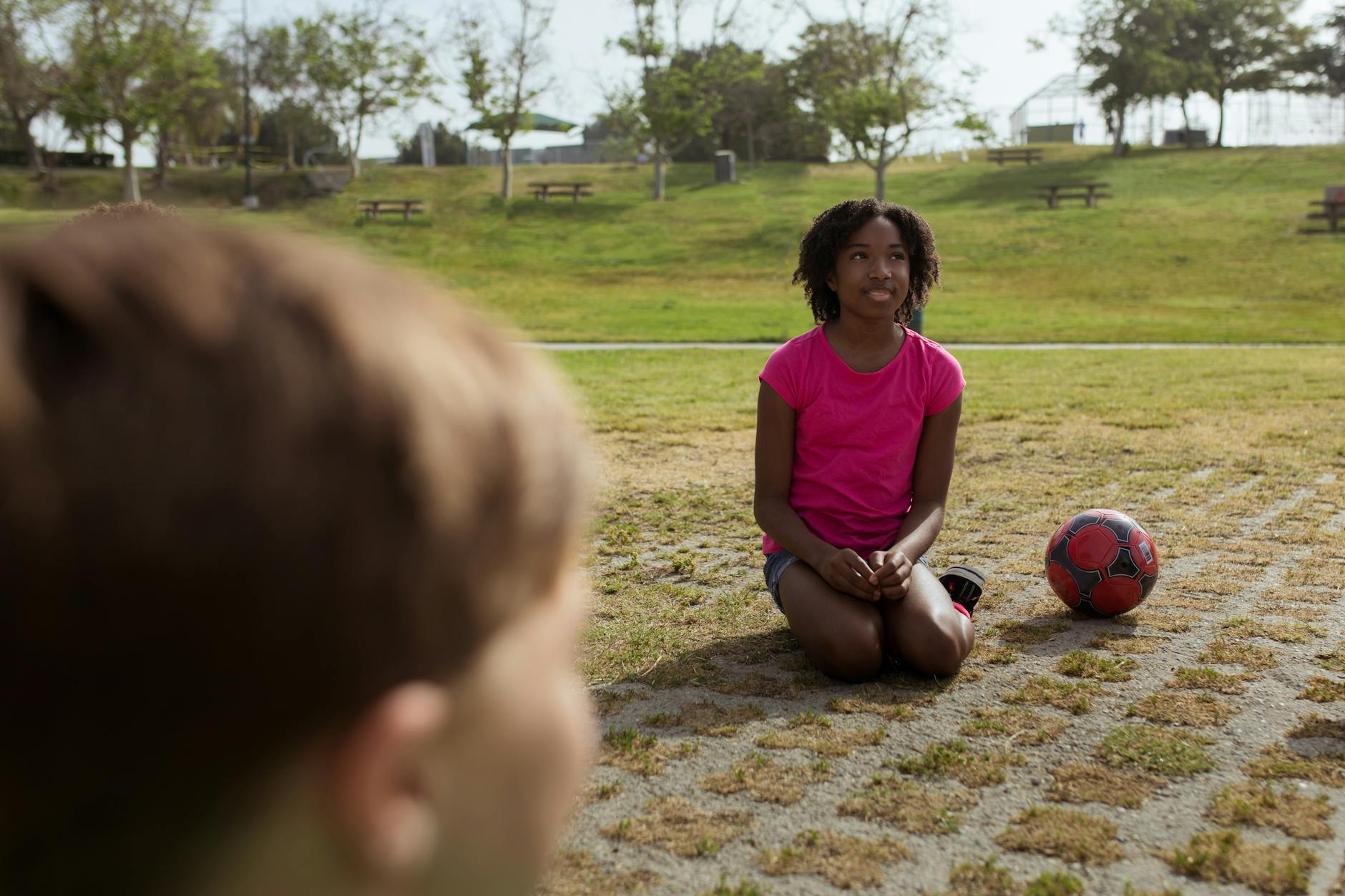 A girl sitting on grass beside a soccer ball in a sunny park, enjoying a playful day.