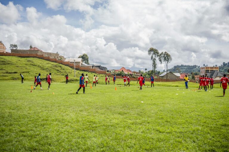 Teenagers playing football on a grassy field with houses and mountains in the background.