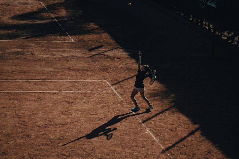 A tennis player serves on a clay court under dramatic lighting, highlighting shadows and motion.
