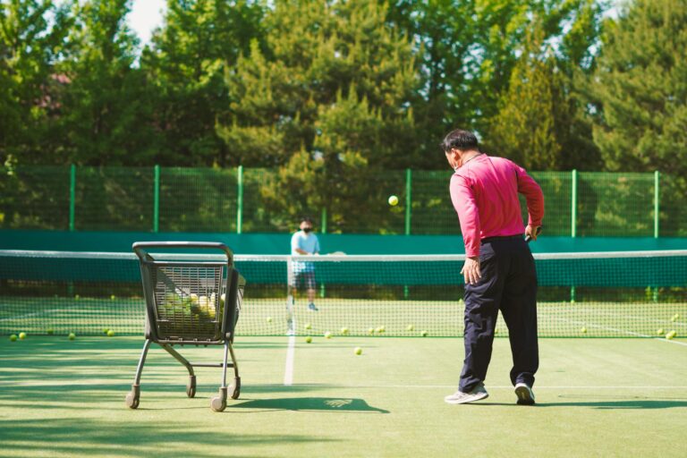 Two men practicing tennis on a sunny outdoor court with a ball cart.