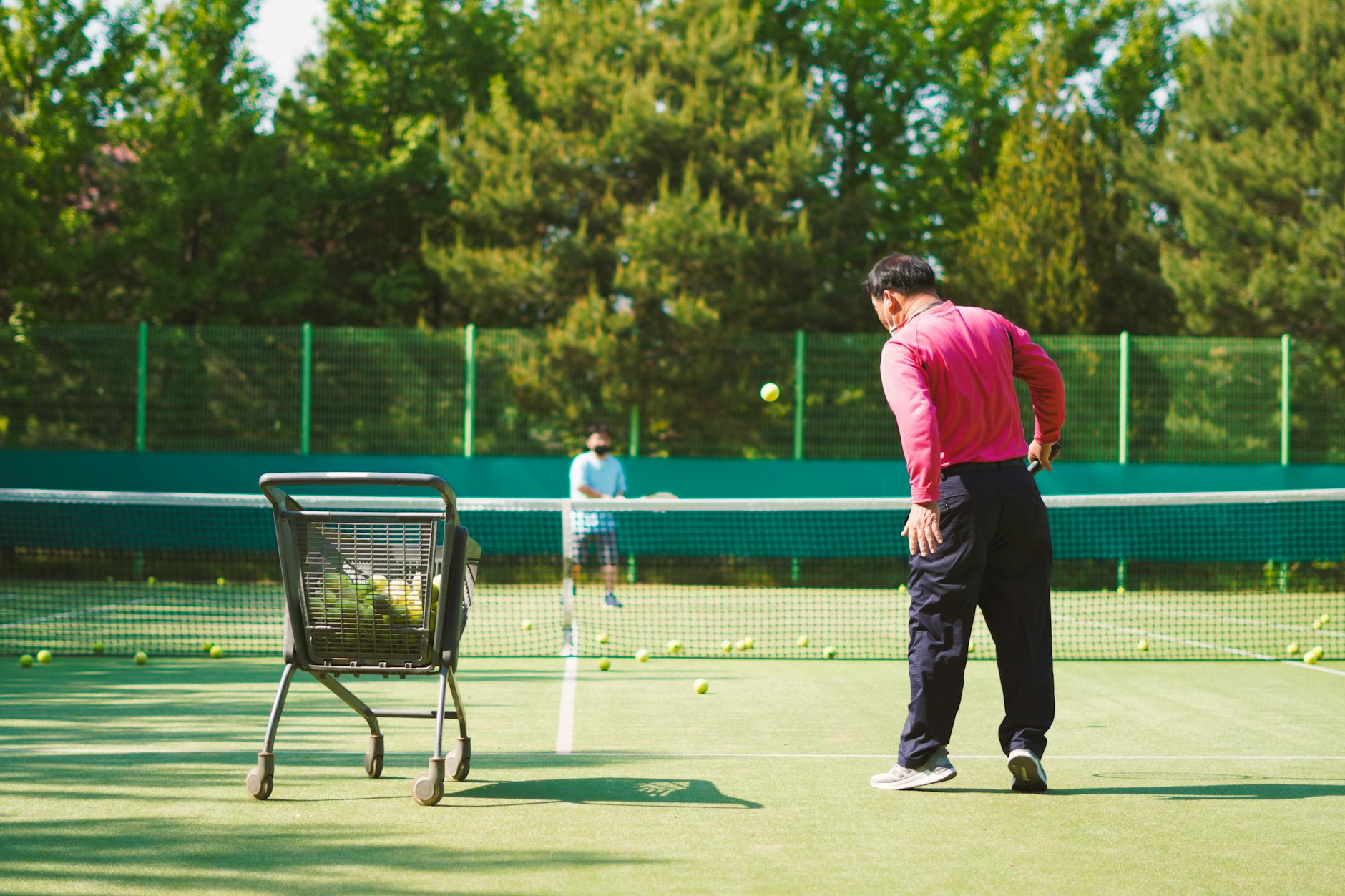 Two men practicing tennis on a sunny outdoor court with a ball cart.