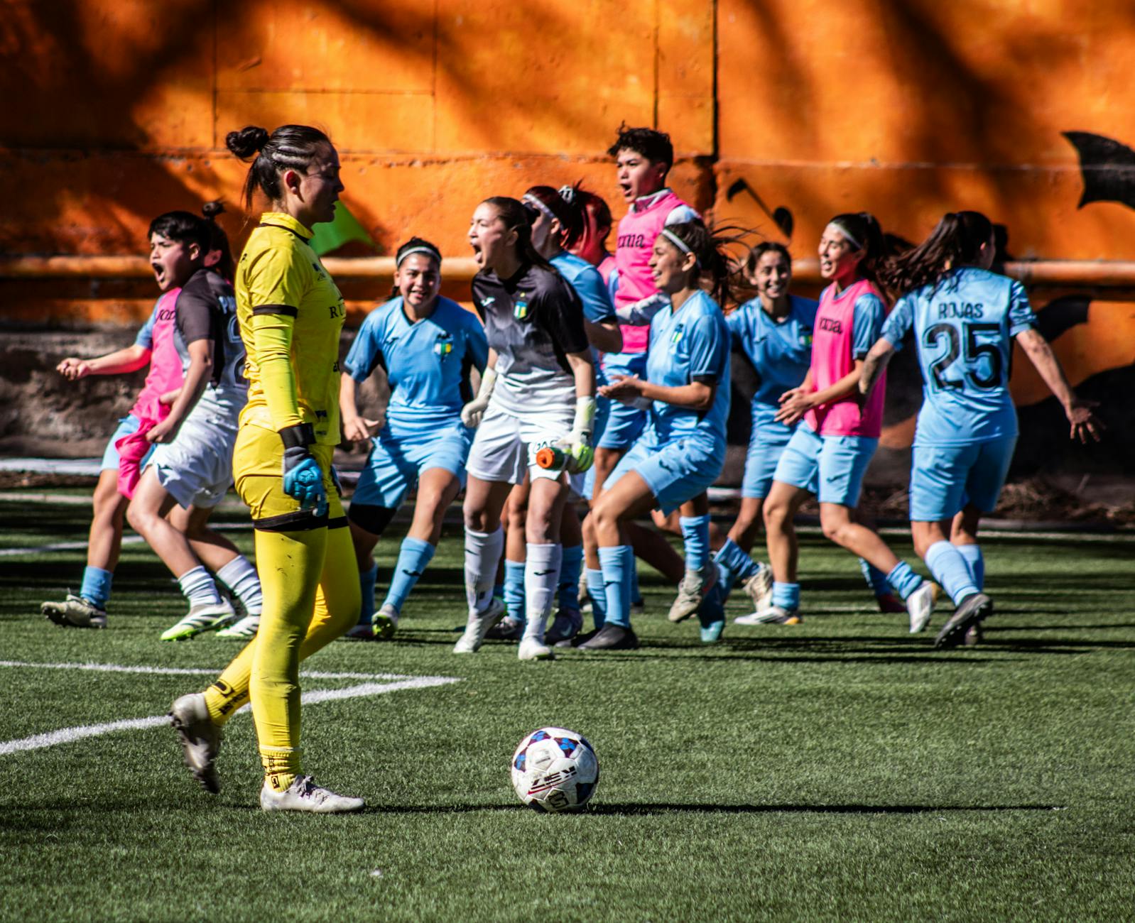 A women's soccer team celebrating after scoring a goal during a daytime match.