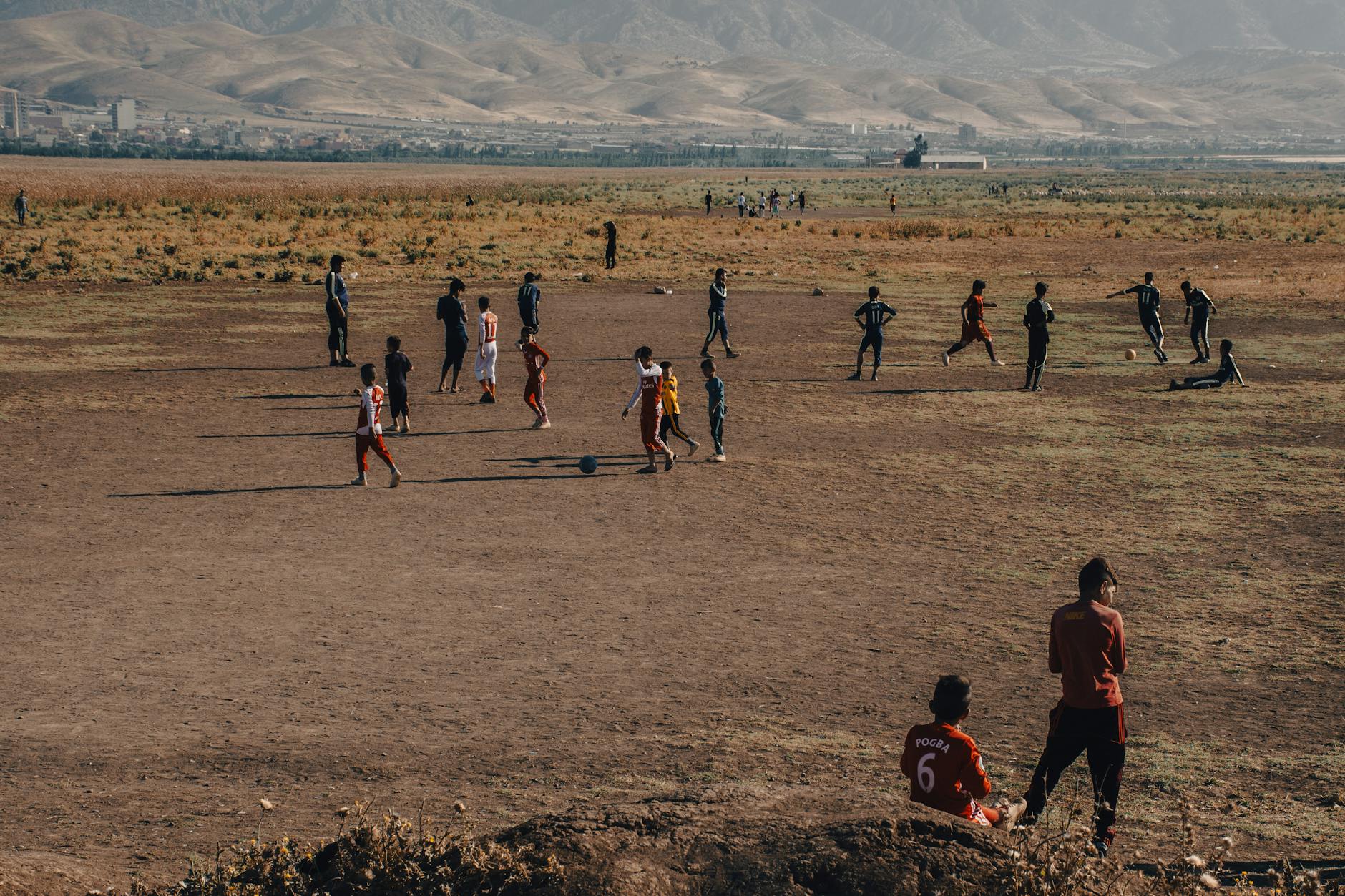A group of children playing soccer in a rural field with mountains in the background. The scene captures a warm, outdoor ambiance.