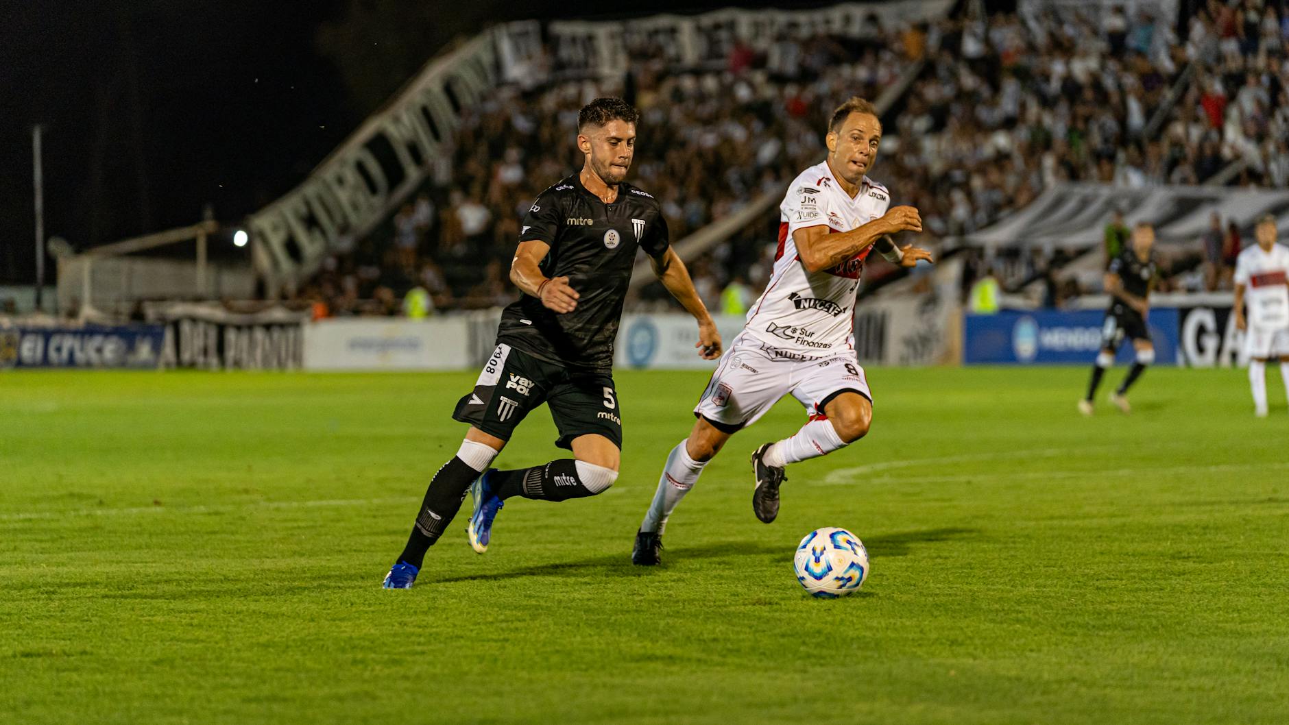 Two soccer players competing vigorously during a night match in a stadium, full crowd in background.